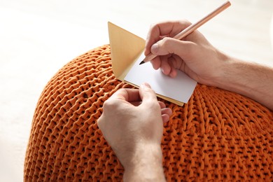 Man writing message in greeting card on orange
knitted pouf, closeup