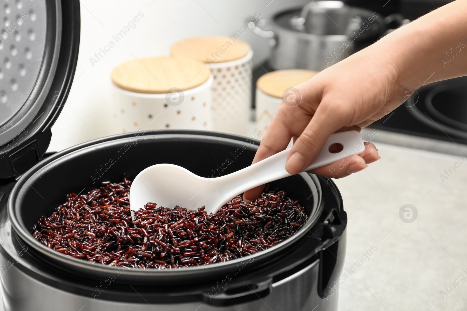 Photo of Woman taking brown rice from multi cooker with spoon in kitchen, closeup