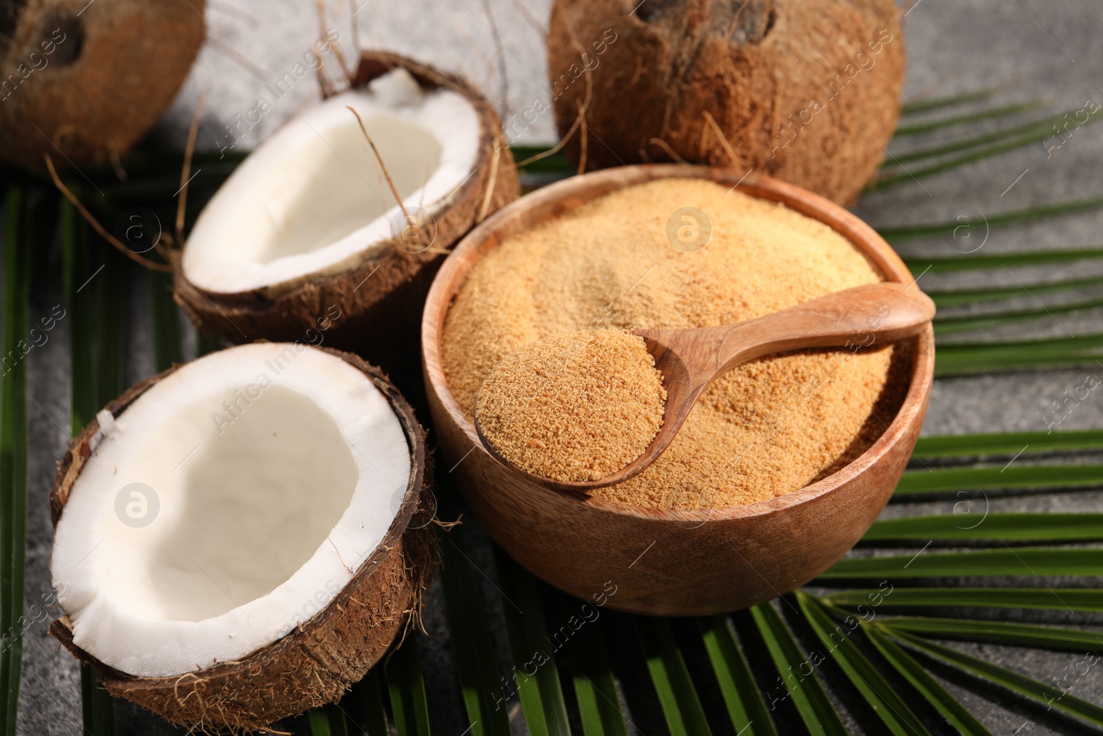 Photo of Spoon with coconut sugar, bowl, palm leaves and fruits on dark textured table, closeup