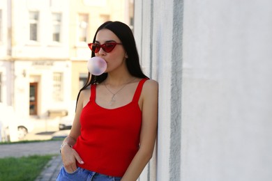 Beautiful woman in stylish sunglasses blowing gum near building outdoors, space for text