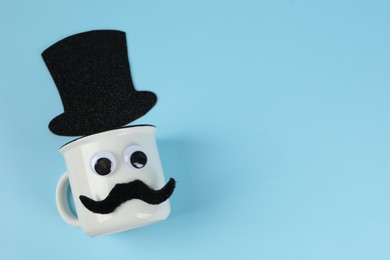 Photo of Man's face made of cup, fake mustache, decorative eyes and hat on light blue background, top view. Space for text