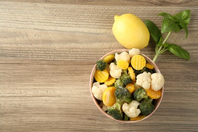 Frozen vegetables in bowl, basil and lemon on wooden table, flat lay. Space for text