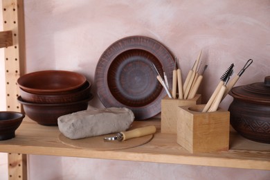 Photo of Set of different crafting tools and clay dishes on wooden rack in workshop