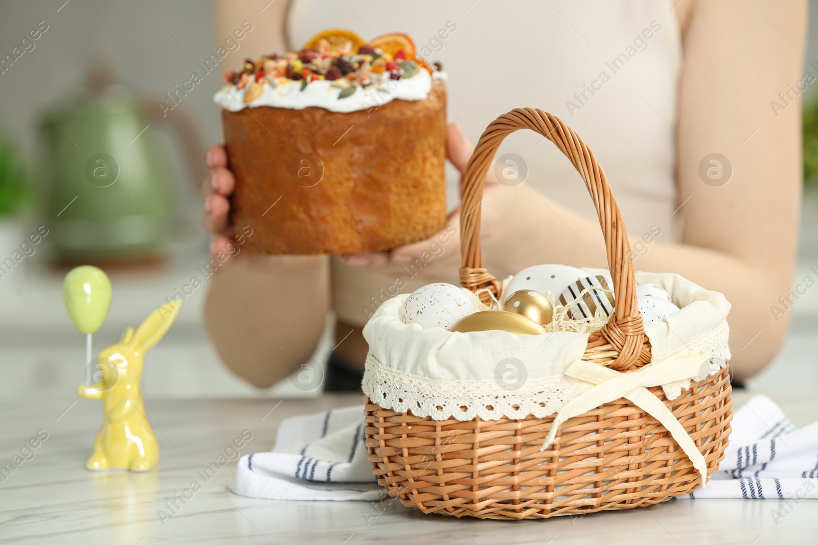 Photo of Closeup of Easter basket with painted eggs on white marble table near woman with cake, focus on food