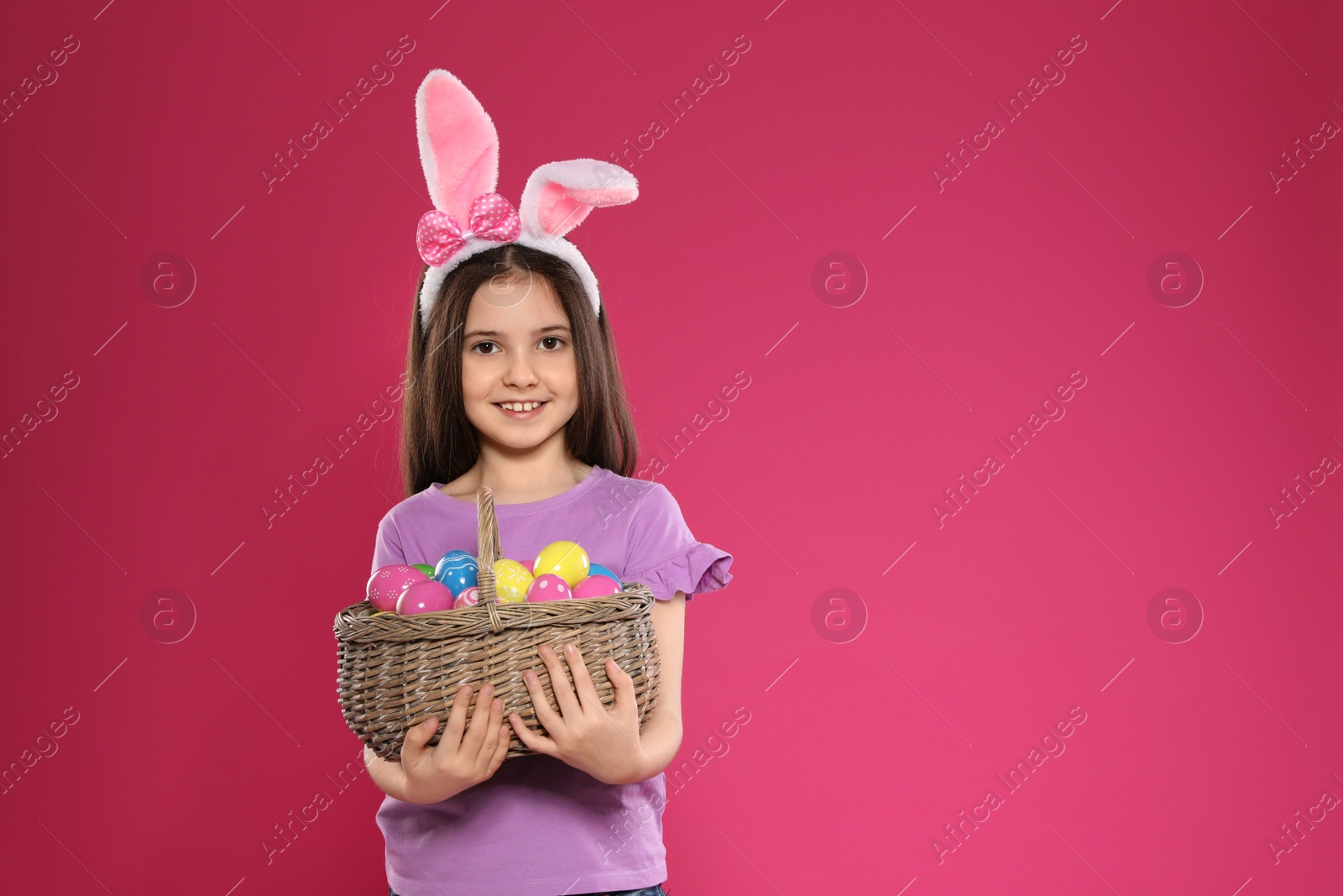 Photo of Little girl in bunny ears headband holding basket with Easter eggs on color background, space for text