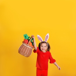 Photo of Happy little girl with bunny ears holding wicker basket full of toy carrots on orange background. Easter celebration