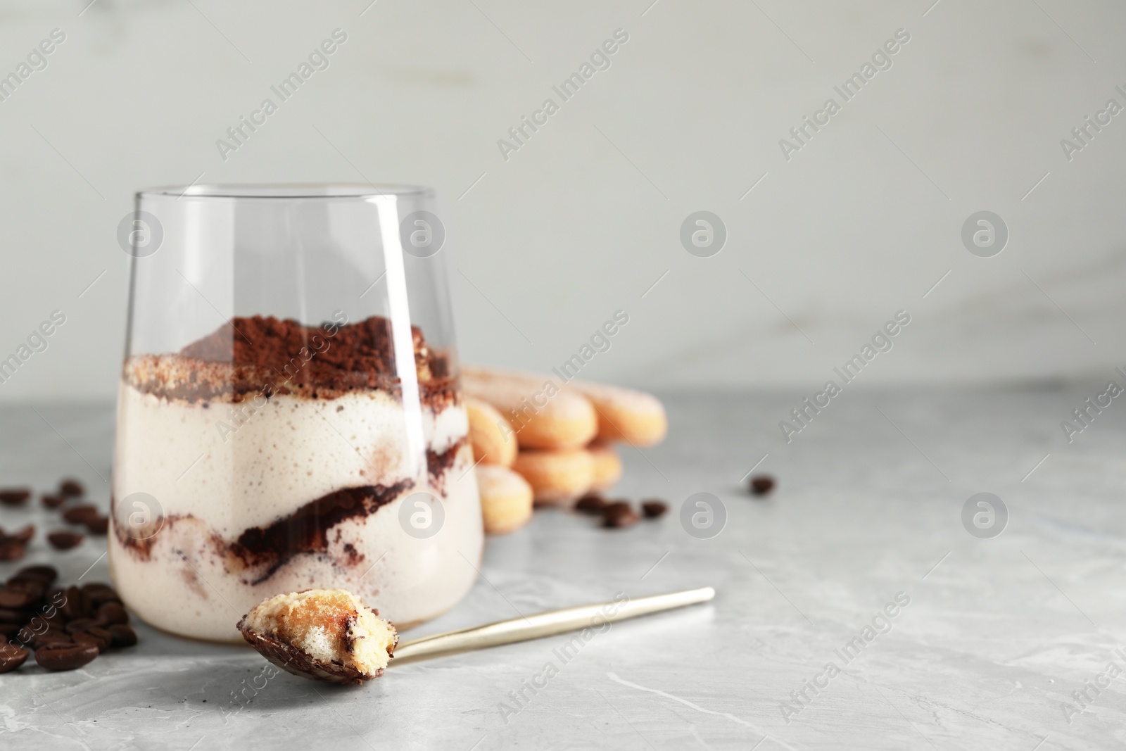 Photo of Tasty tiramisu in glass, coffee beans, spoon and biscuits on light grey table, closeup. Space for text