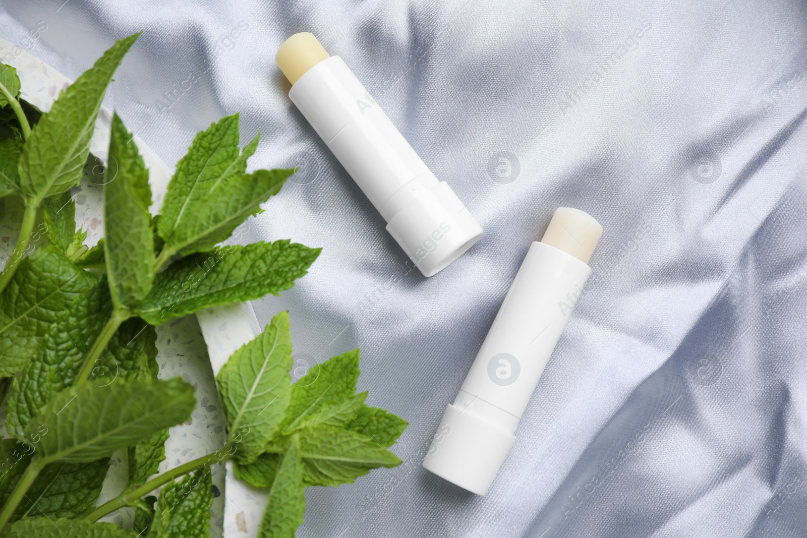 Photo of Hygienic lipsticks and mint leaves on light silk fabric, flat lay