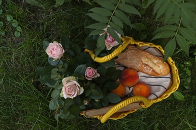Yellow wicker bag with peaches, roses, book and baguette on green grass outdoors, top view
