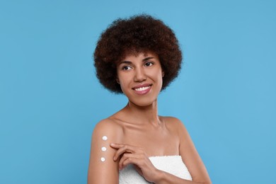 Beautiful young woman applying body cream onto arm on light blue background