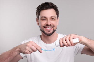 Photo of Happy man squeezing toothpaste from tube onto toothbrush on light grey background