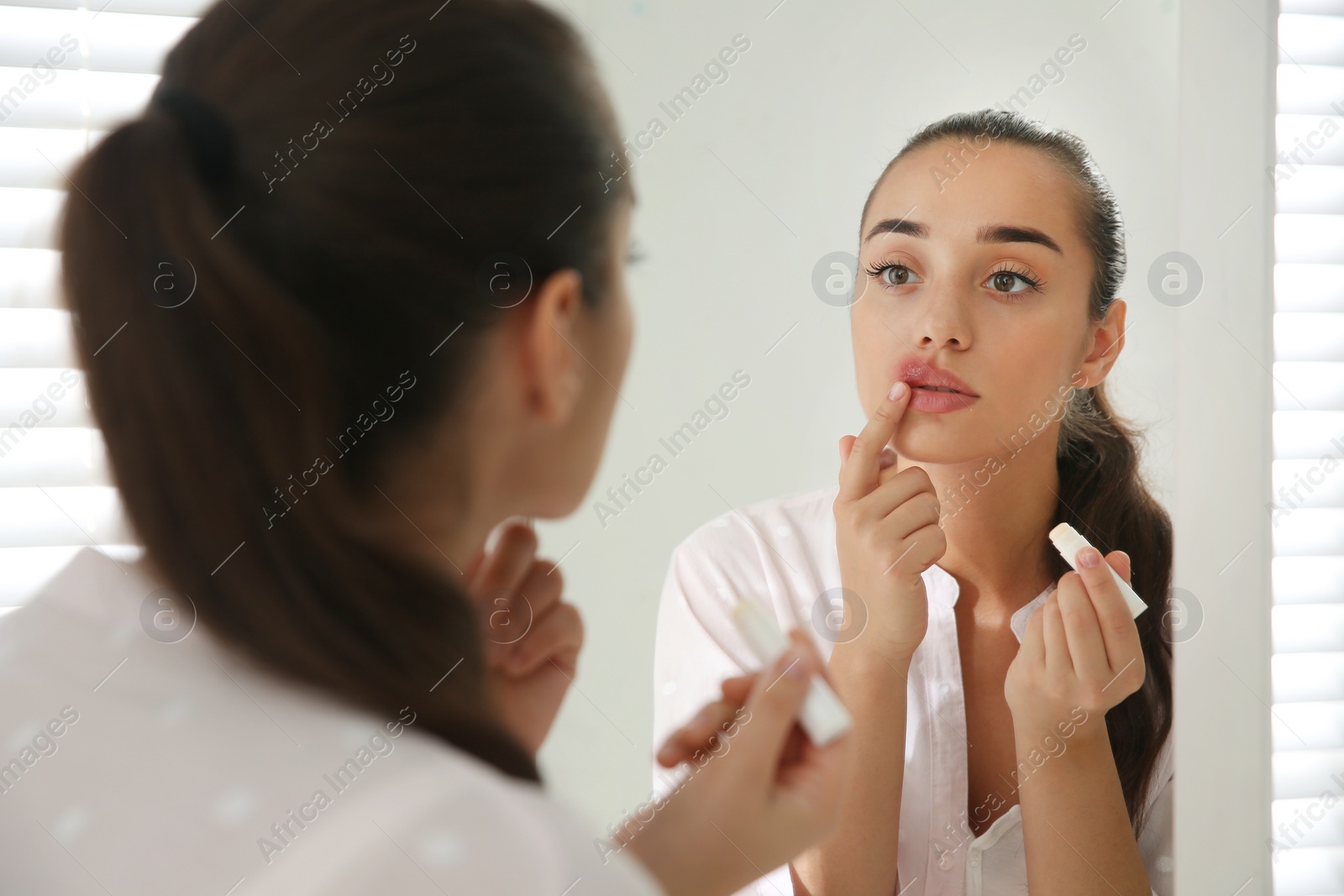 Photo of Young woman with herpes applying lip balm in front of mirror at home