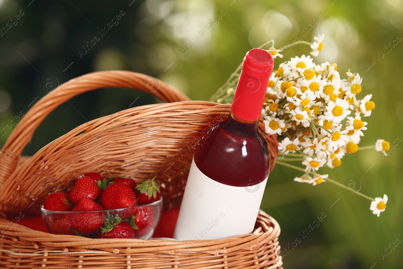 Photo of Picnic basket with wine, strawberries and flowers on blurred background, closeup