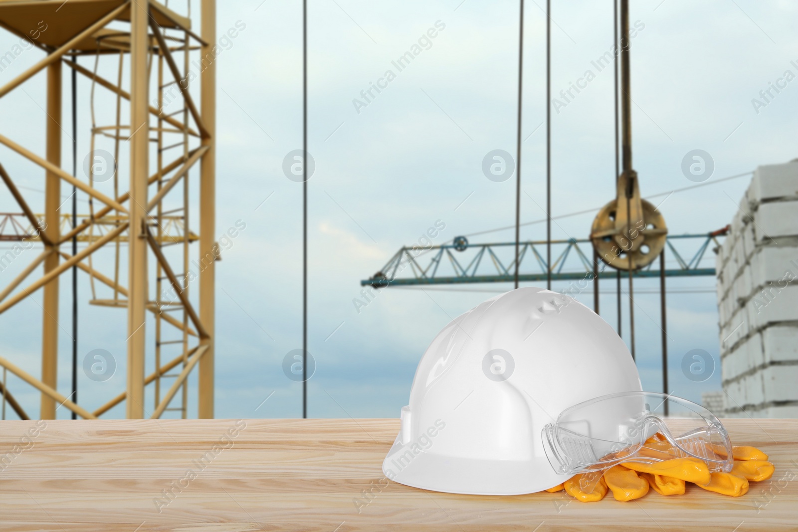 Image of Safety equipment. Hard hat, gloves and goggles on wooden surface near construction site with tower crane outdoors, space for text