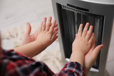 Photo of Mother with her kid warming hands near electric heater at home, closeup