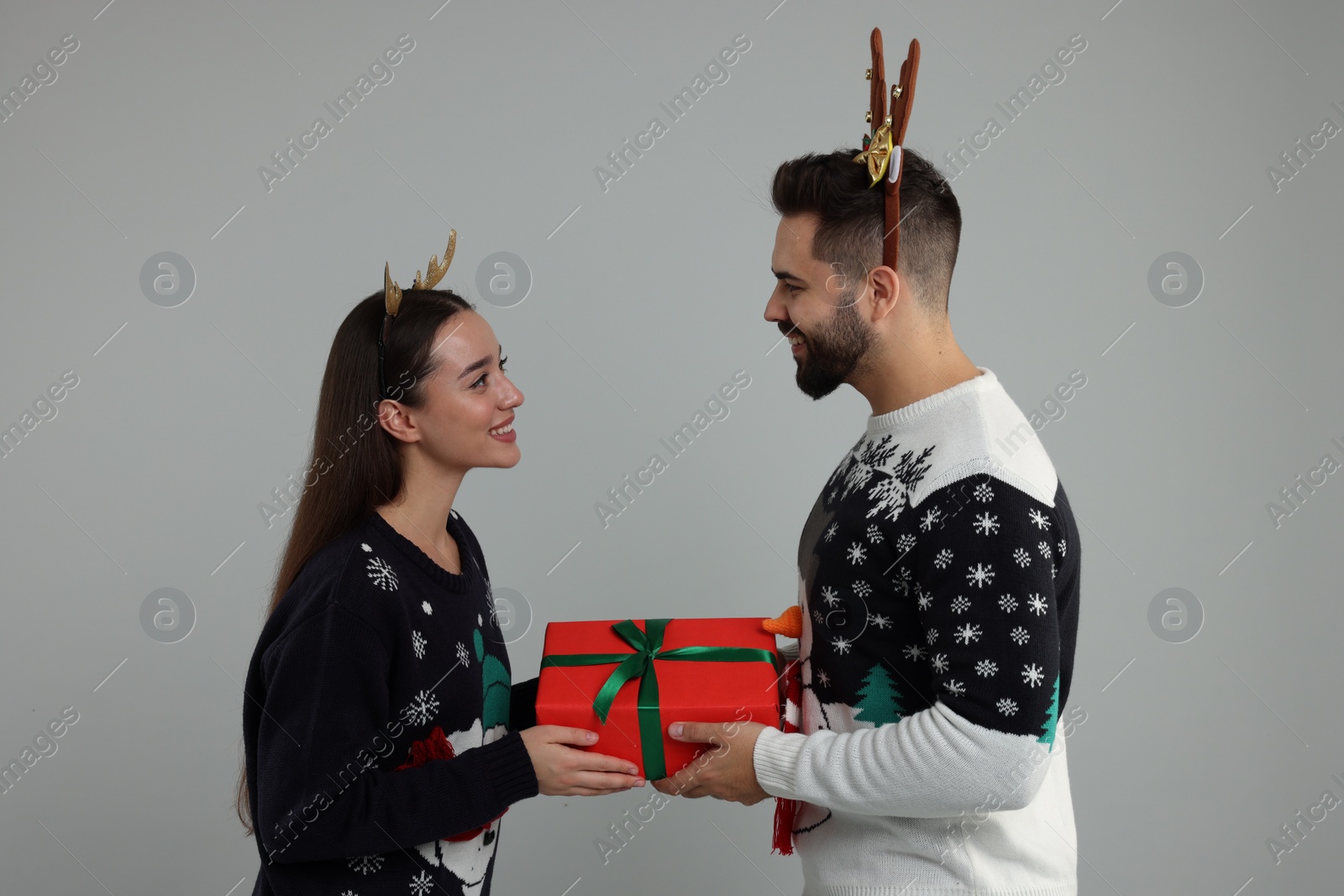 Photo of Happy young couple in Christmas sweaters and reindeer headbands holding gift on grey background