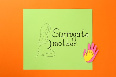 Photo of Paper note with drawn pregnant woman figure and words Surrogate mother on orange background, top view