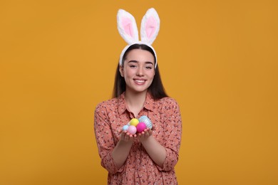 Photo of Happy woman in bunny ears headband holding painted Easter eggs on orange background