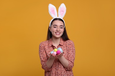 Happy woman in bunny ears headband holding painted Easter eggs on orange background