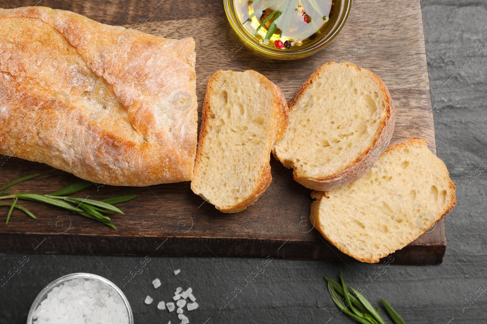 Photo of Delicious French baguette with rosemary, oil and salt on black table, flat lay