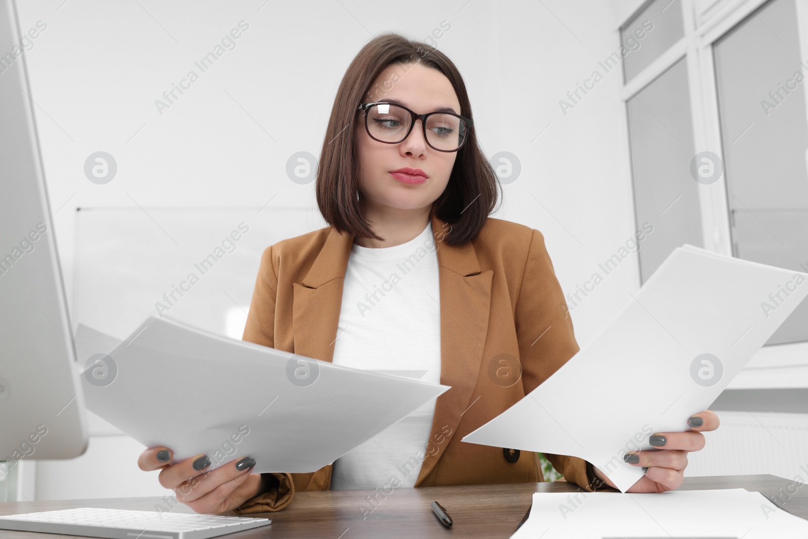 Photo of Puzzled young intern with paper sheets at table in modern office. First work day