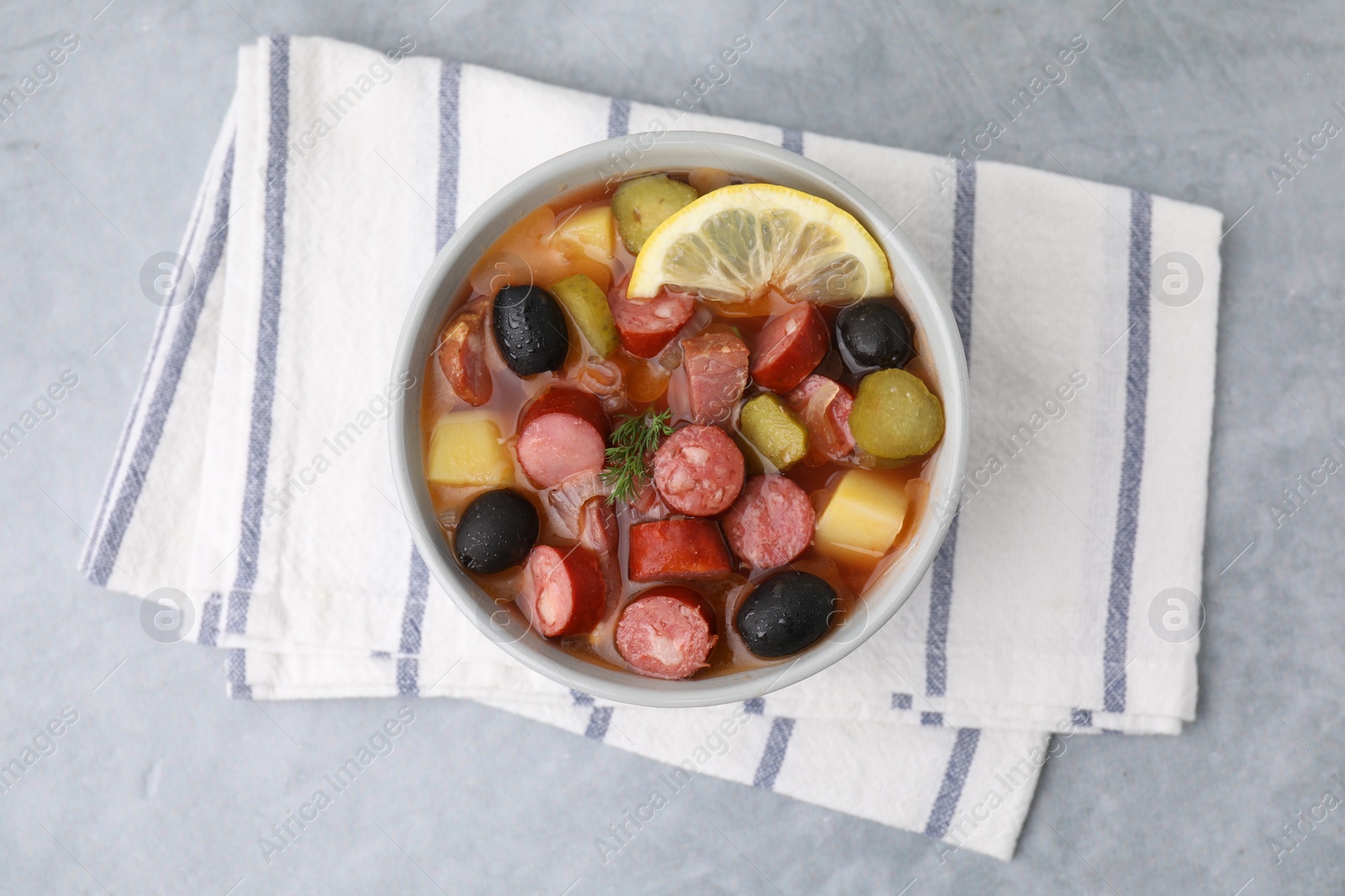 Photo of Meat solyanka soup with thin dry smoked sausages in bowl on grey table, top view