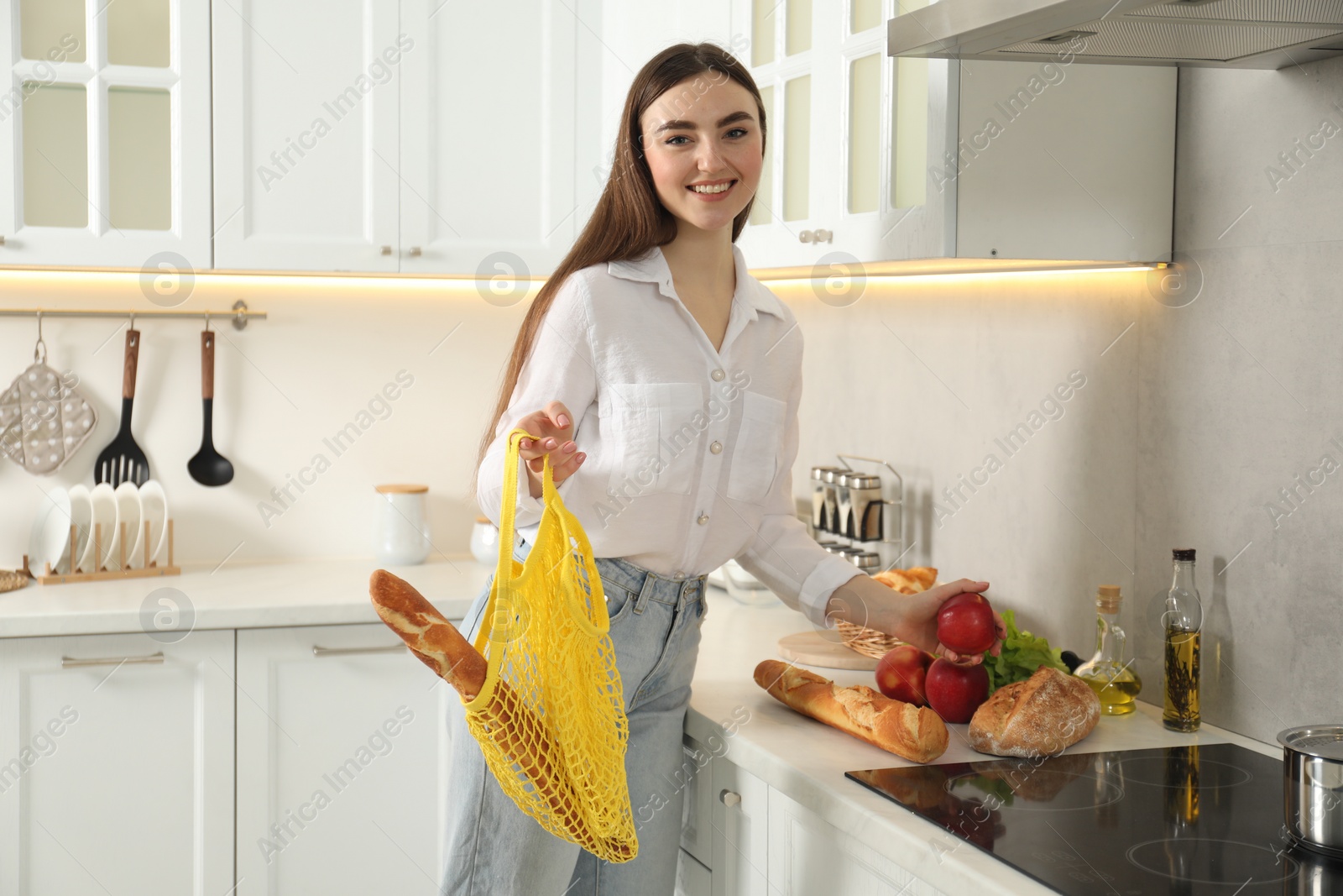 Photo of Woman holding string bag with baguette and apple in kitchen