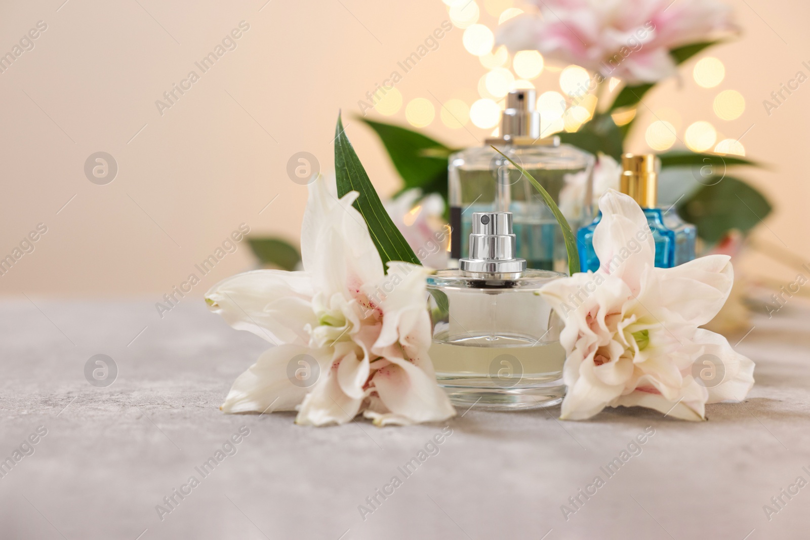 Photo of Bottles of perfume and beautiful lily flowers on table against beige background with blurred lights, closeup. Space for text