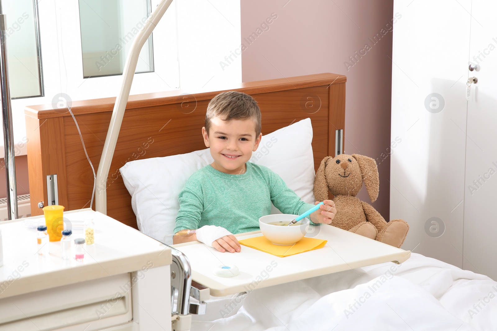 Photo of Little child with intravenous drip eating soup in hospital bed