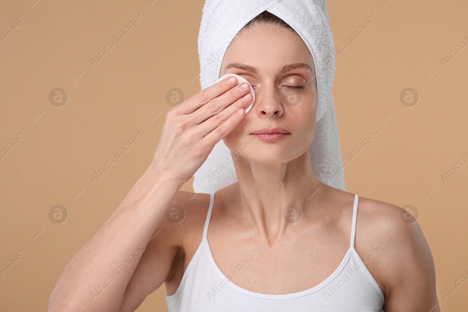 Photo of Beautiful woman in terry towel removing makeup with cotton pad on beige background