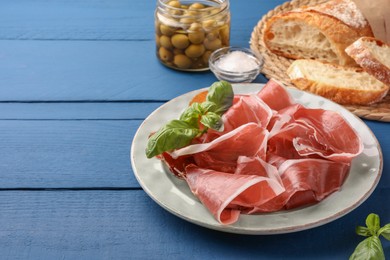 Slices of tasty cured ham, basil, bread and olives on blue wooden table. Space for text