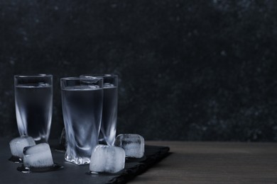 Photo of Shot glasses of vodka with ice cubes on wooden table against black wall. Space for text