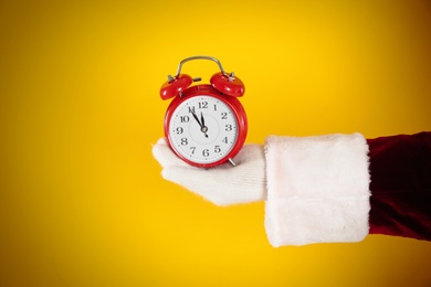 Photo of Santa Claus holding alarm clock on yellow background, closeup. Christmas countdown