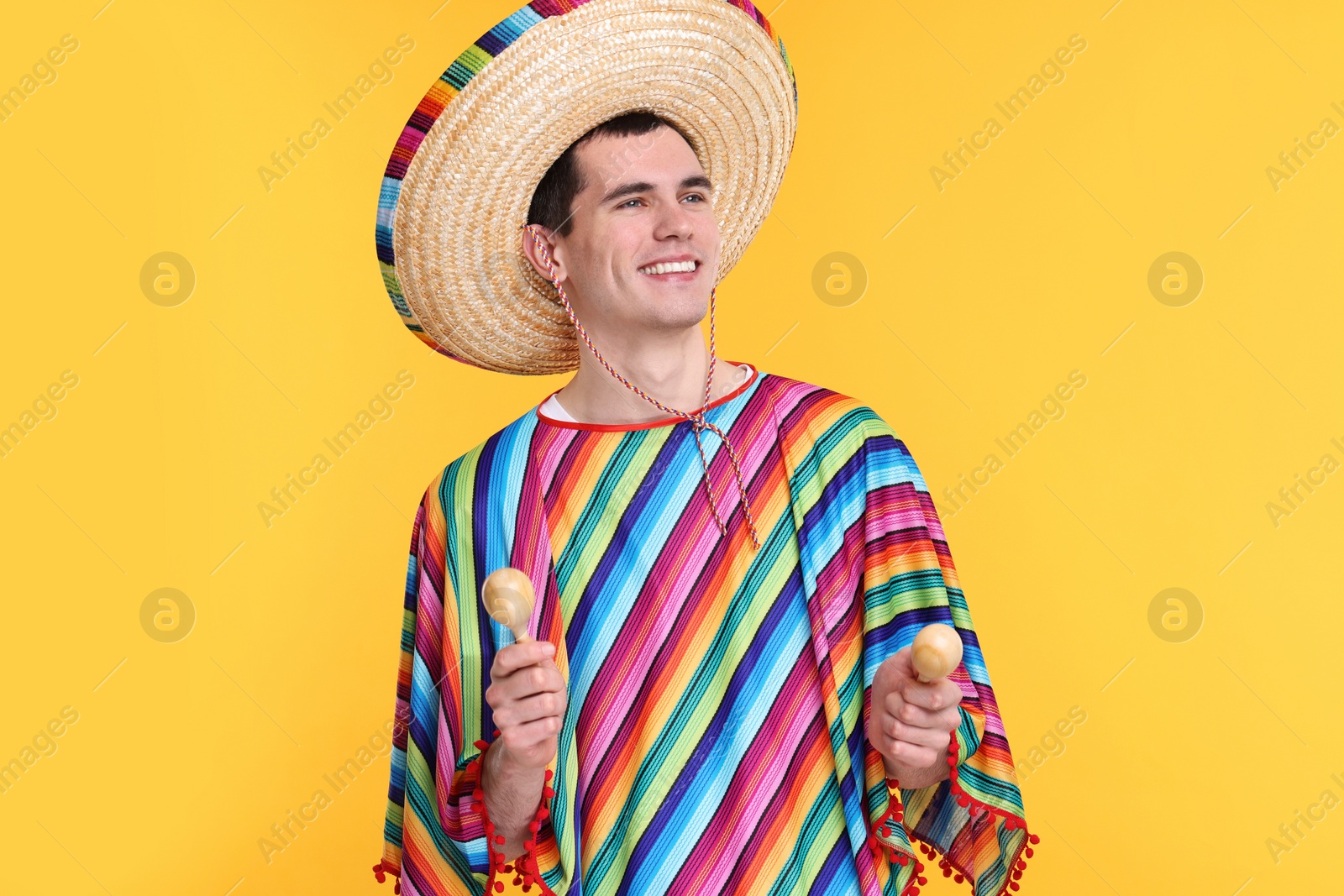 Photo of Young man in Mexican sombrero hat and poncho with maracas on yellow background