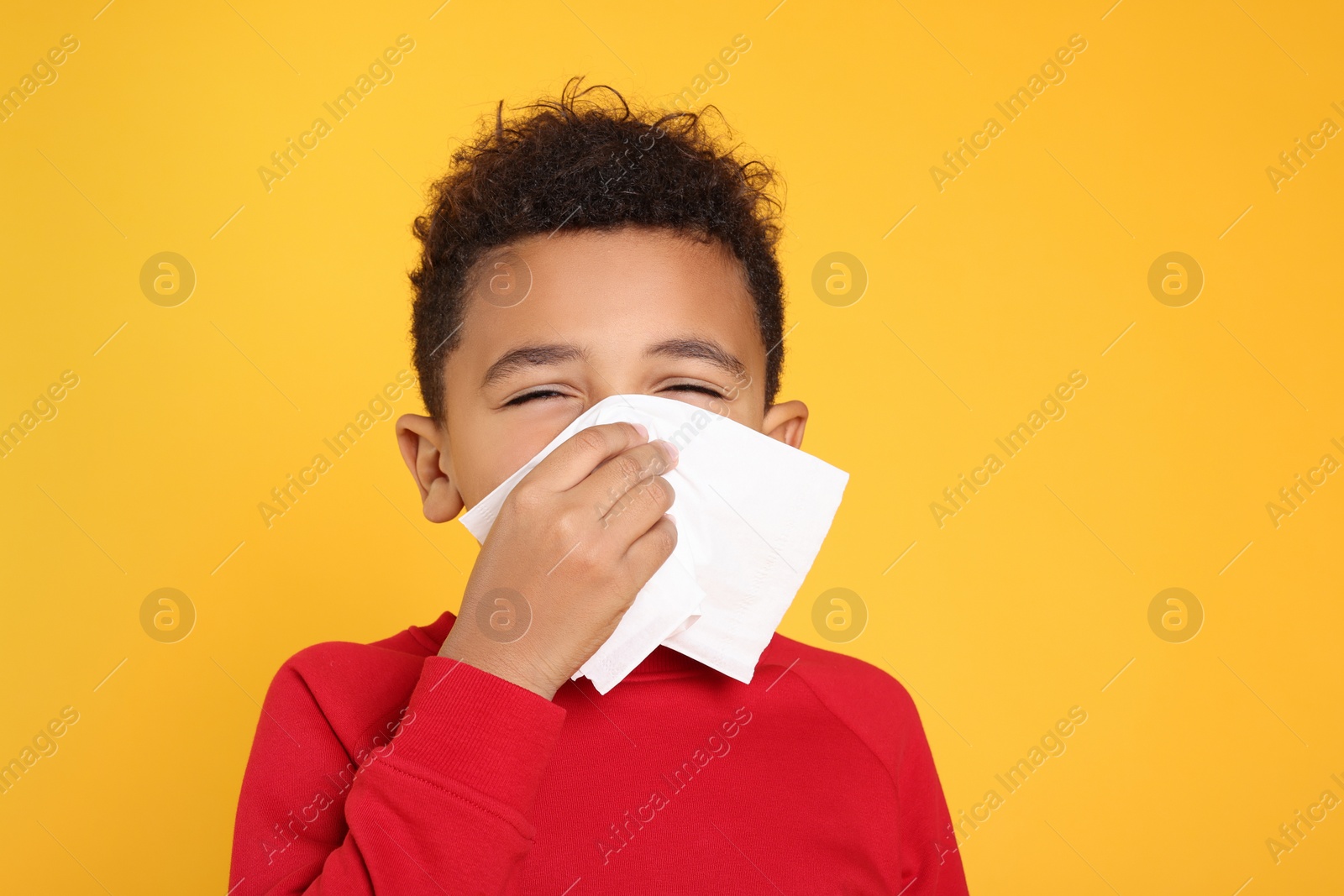 Photo of African-American boy blowing nose in tissue on yellow background. Cold symptoms