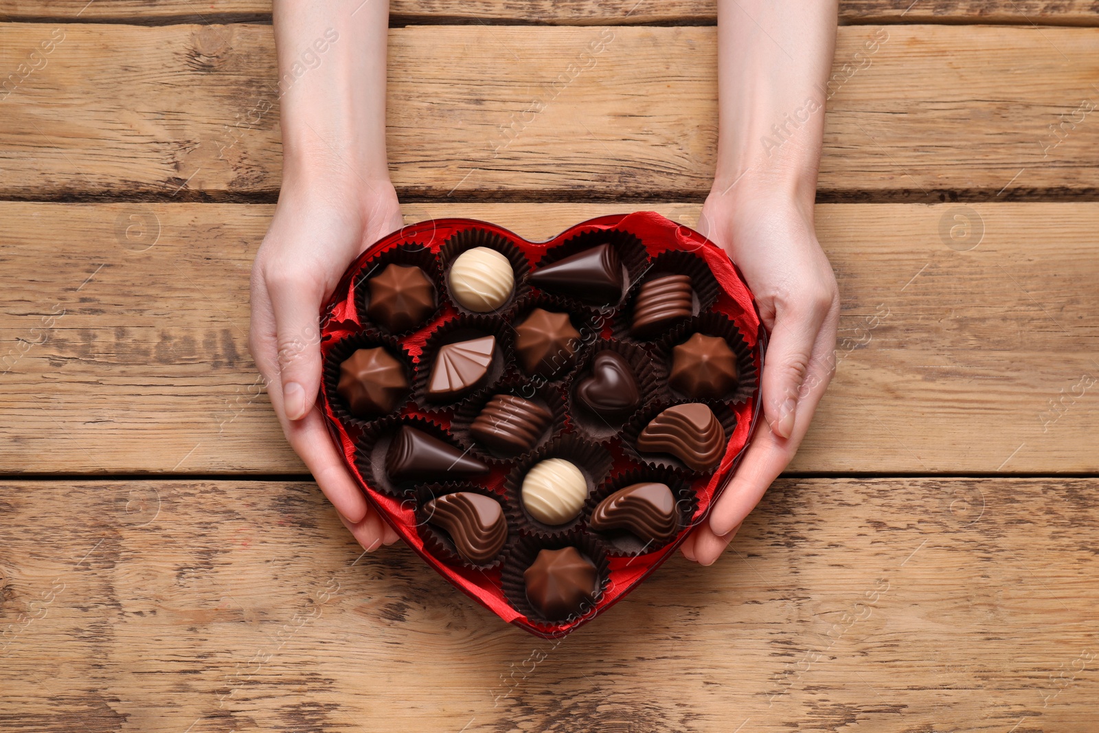 Photo of Woman holding heart shaped box with delicious chocolate candies at wooden table, top view