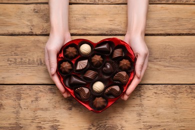 Woman holding heart shaped box with delicious chocolate candies at wooden table, top view