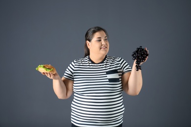 Overweight woman with grapes and hamburger on gray background
