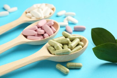 Photo of Different vitamin pills in spoons and green leaves on light blue background, closeup