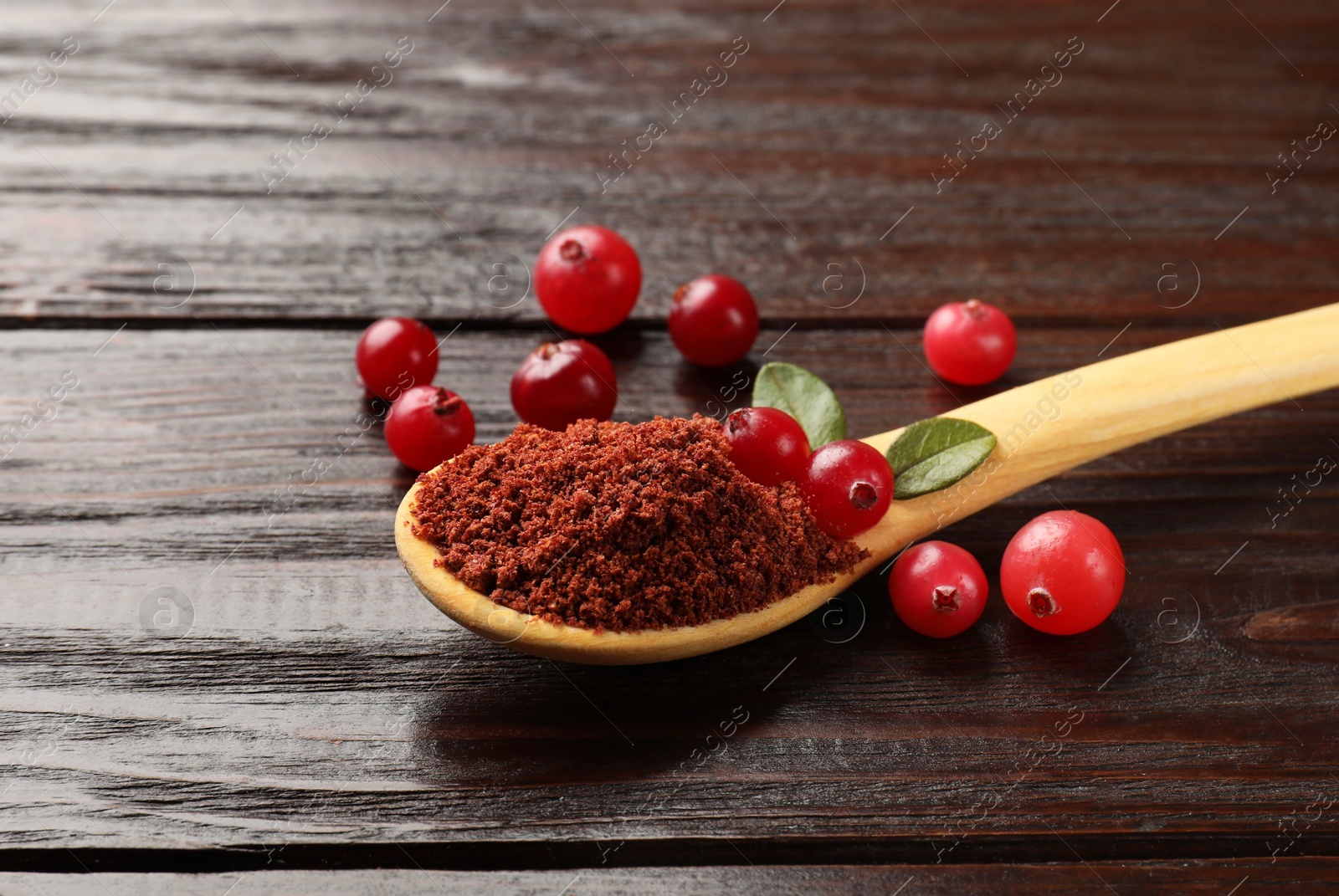 Photo of Dried cranberry powder in spoon, fresh berries and green leaves on wooden table, closeup