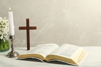 Burning church candle, Bible, wooden cross and flowers on white table