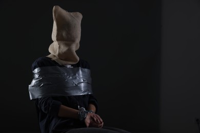 Taped up woman with bag on her head on dark background, space for text. Hostage