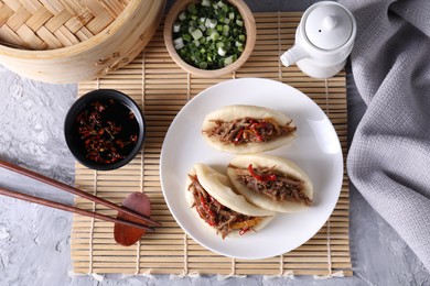 Photo of Delicious gua bao (pork belly buns), chopsticks, green onion and sauces on grey textured table, flat lay