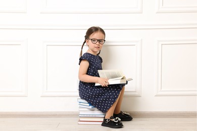 Photo of Cute little girl in glasses sitting on stack of books near white wall