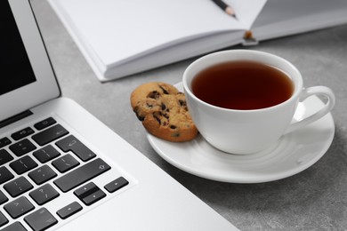 Photo of Chocolate chip cookie, cup of tea and laptop on light grey table, closeup