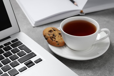 Chocolate chip cookie, cup of tea and laptop on light grey table, closeup