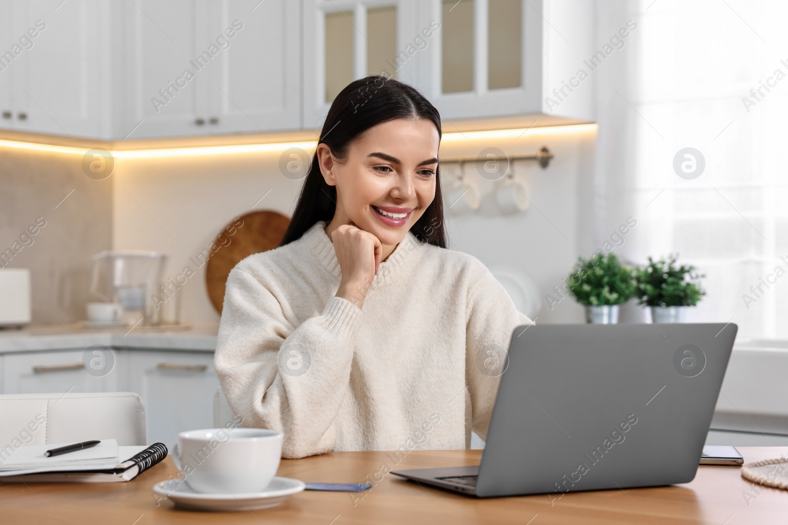 Photo of Happy young woman with laptop shopping online at wooden table in kitchen