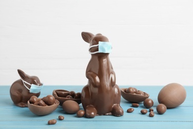 Photo of Chocolate bunnies with protective masks and eggs on light blue wooden table. Easter holiday during COVID-19 quarantine