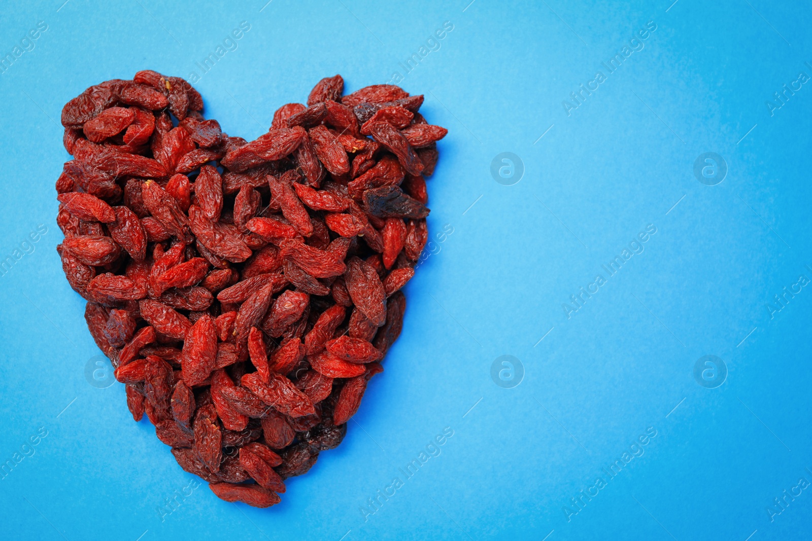 Photo of Heart shaped pile of dried goji berries on light blue background, top view. Space for text