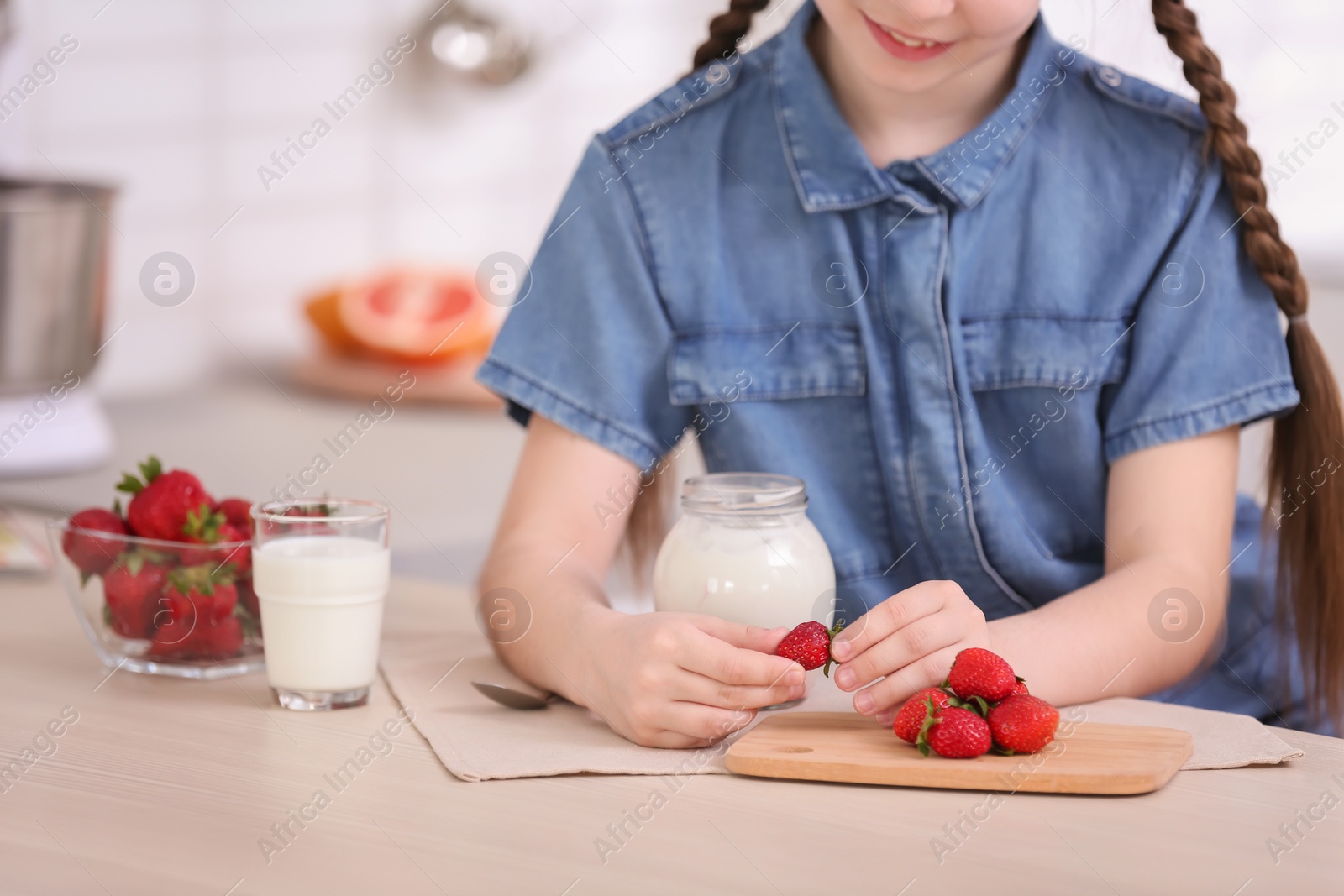 Photo of Cute girl adding strawberry to yogurt in jar at table